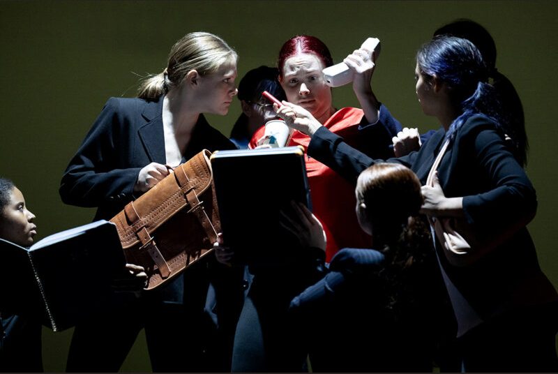 Dancers surround one performer, while holding a book, briefcase and phone toward her face.