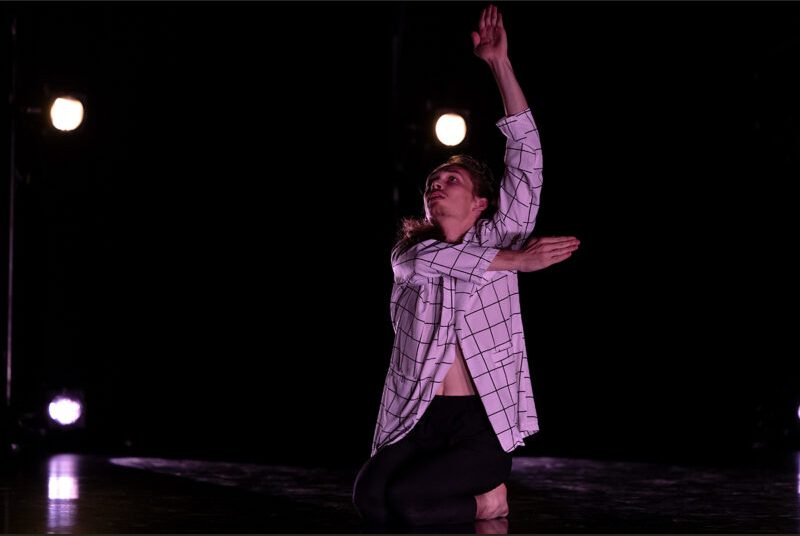 A dancer performs with his left arm stretched upward and his right pointing back toward the left.