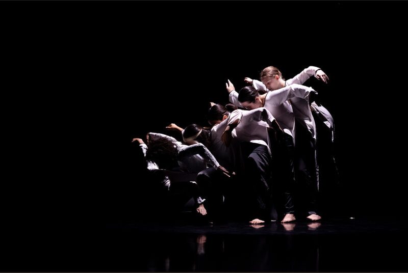 Six dancers stand close together with arms outstretched during a performance.
