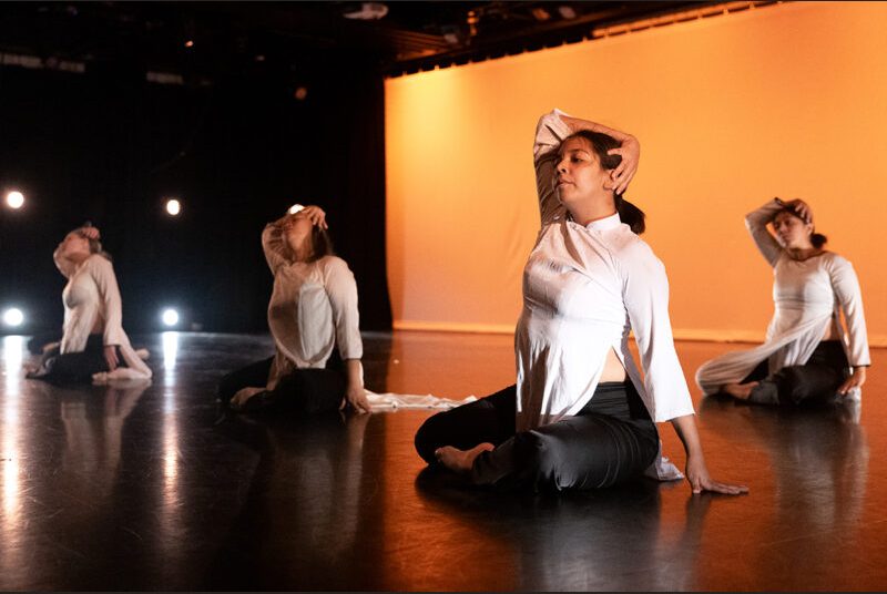 Four dancers sit on the floor during a performance, each placing their right arm on top of their head.