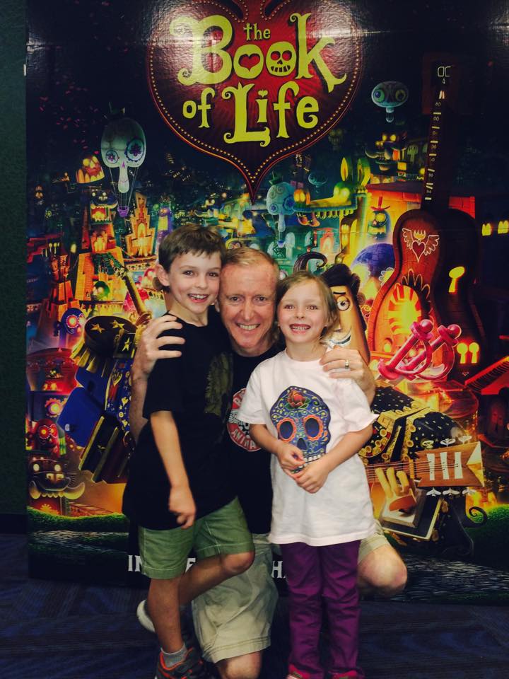 A father poses with his son and daughter in front of a movie poster with the title "The Book Of Life."