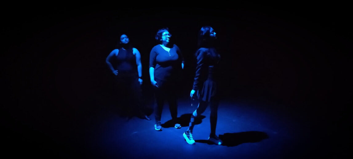 Three college students perform an immersive theatre show with a blue light surrounding them.