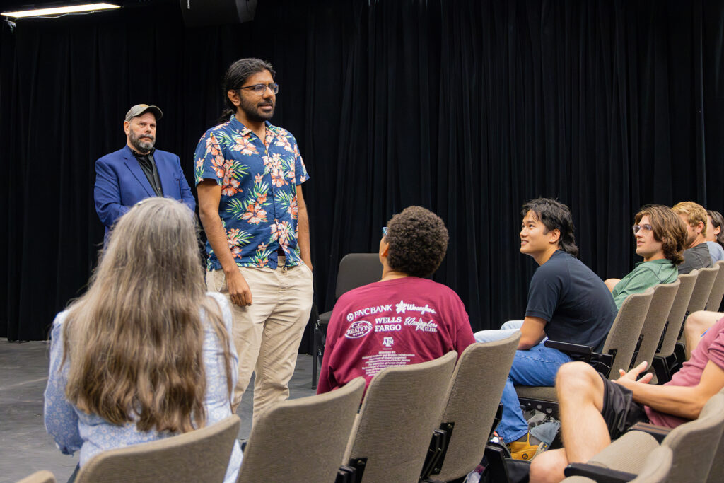 A college student stands in a theater at an improv-comedy workshop, speaking in front of an audience. A man stands behind him.