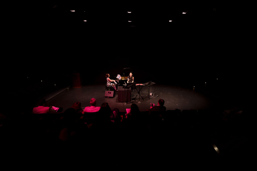 Two performers play piano and toy piano in a theater.