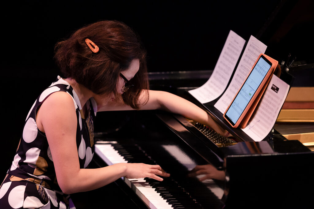A performer plays piano, with her right hand on the keys and her left working with a string on the side opposite of the keys.