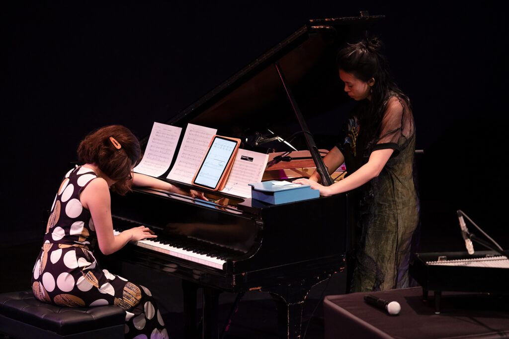 A piano player sits as she performs, while a standing performer plays a toy piano.