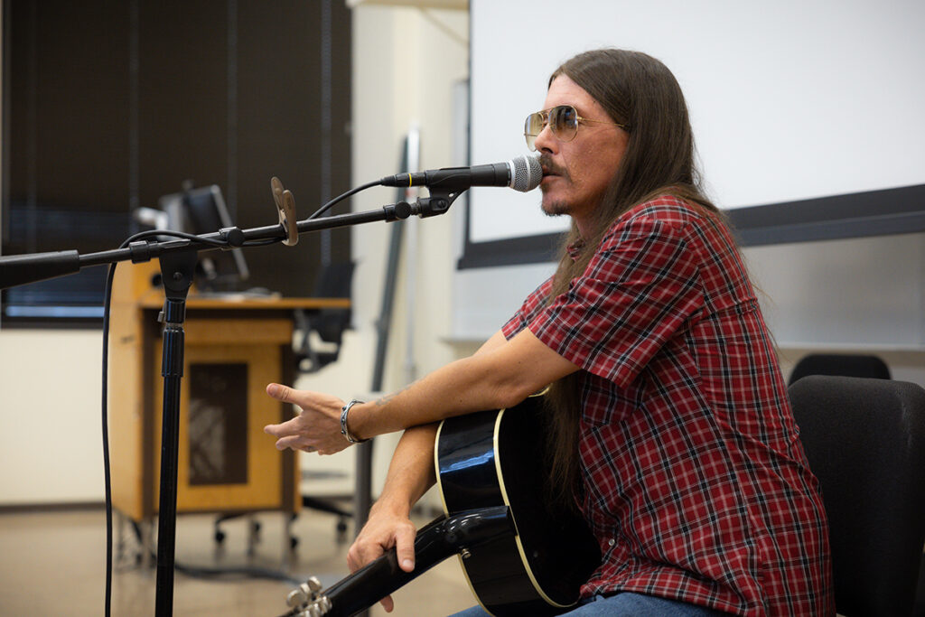 A singer-songwriter smiles while giving a performance for college students in a classroom.