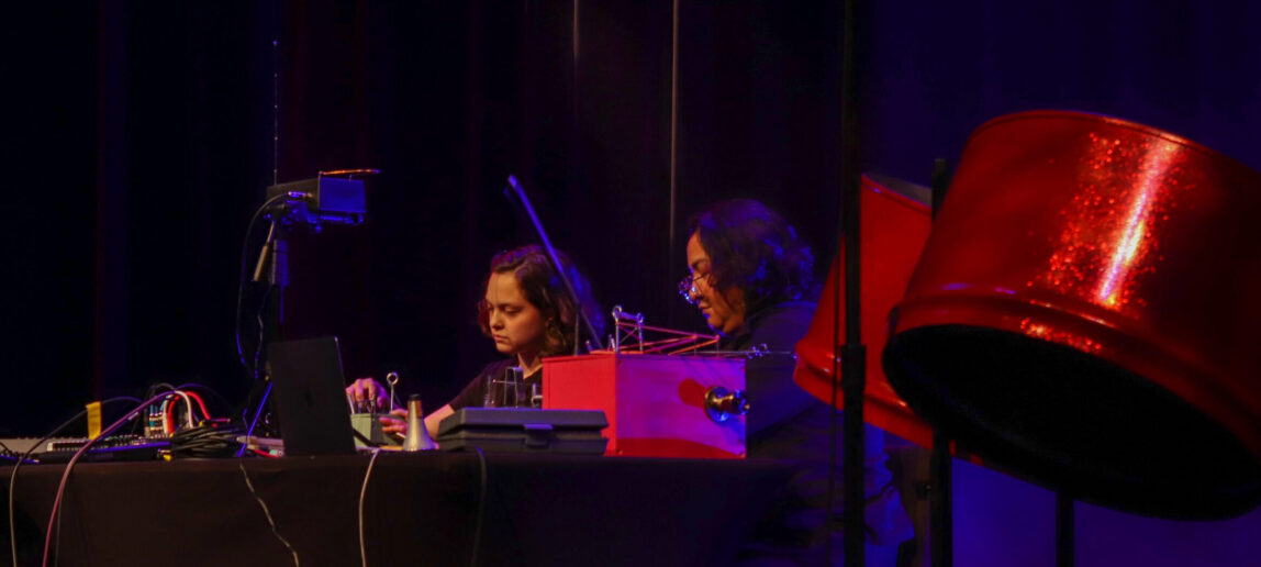 Two college students are seated at a table onstage, performing by using analogue equipment.