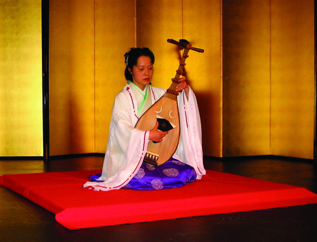 A woman sits on a red mat and plays a traditional Japanese instrument.