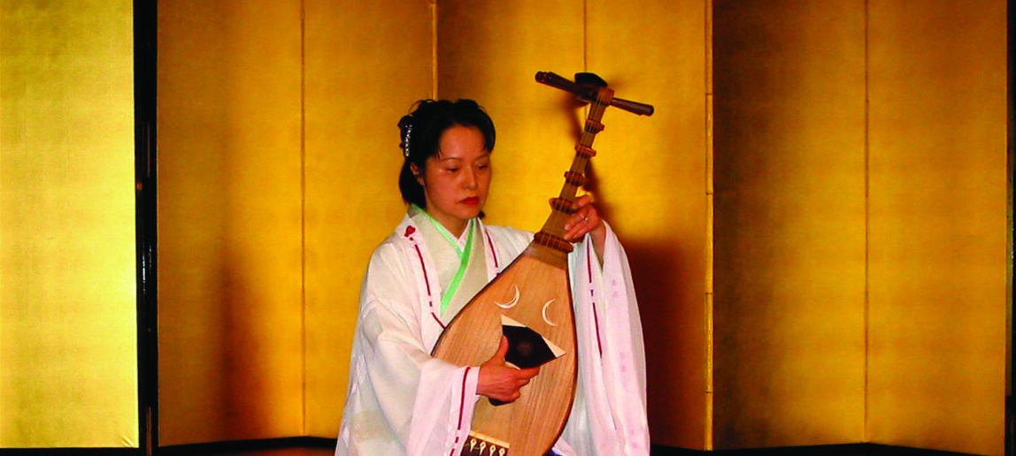 A woman sits on a red mat and plays a traditional Japanese instrument.