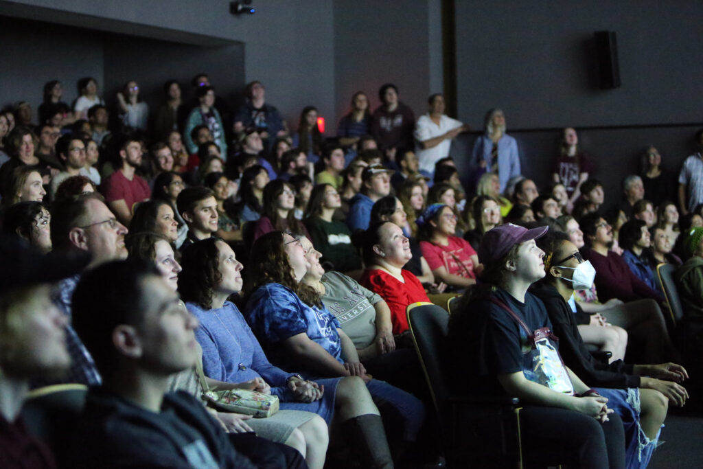 A crowded theater full of college students and professors looks up at a movie screen.