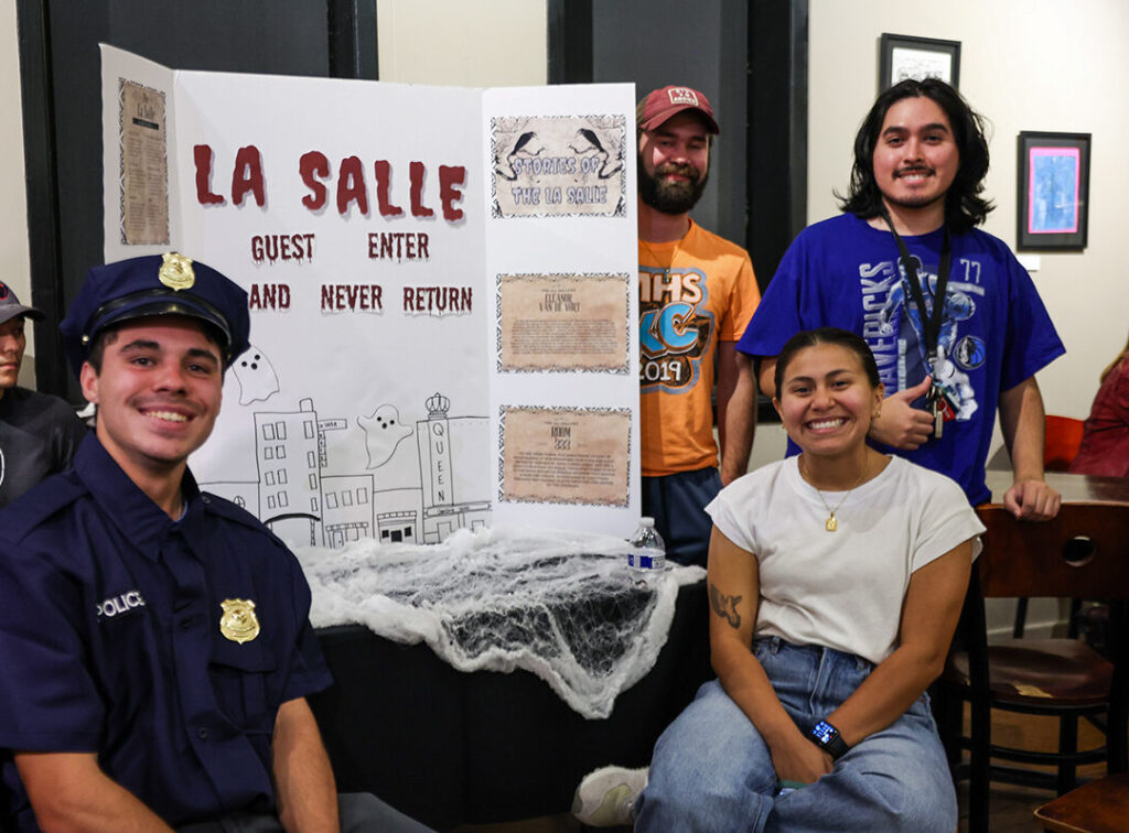 A group of college students pose around a presentation board with drawings and clippings that reads: "La Salle. Guest enter and never return."