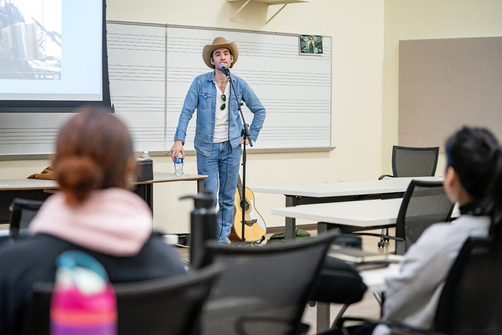 A country music performer stands in front of a microphone stand as he speaks with college students in a classroom.