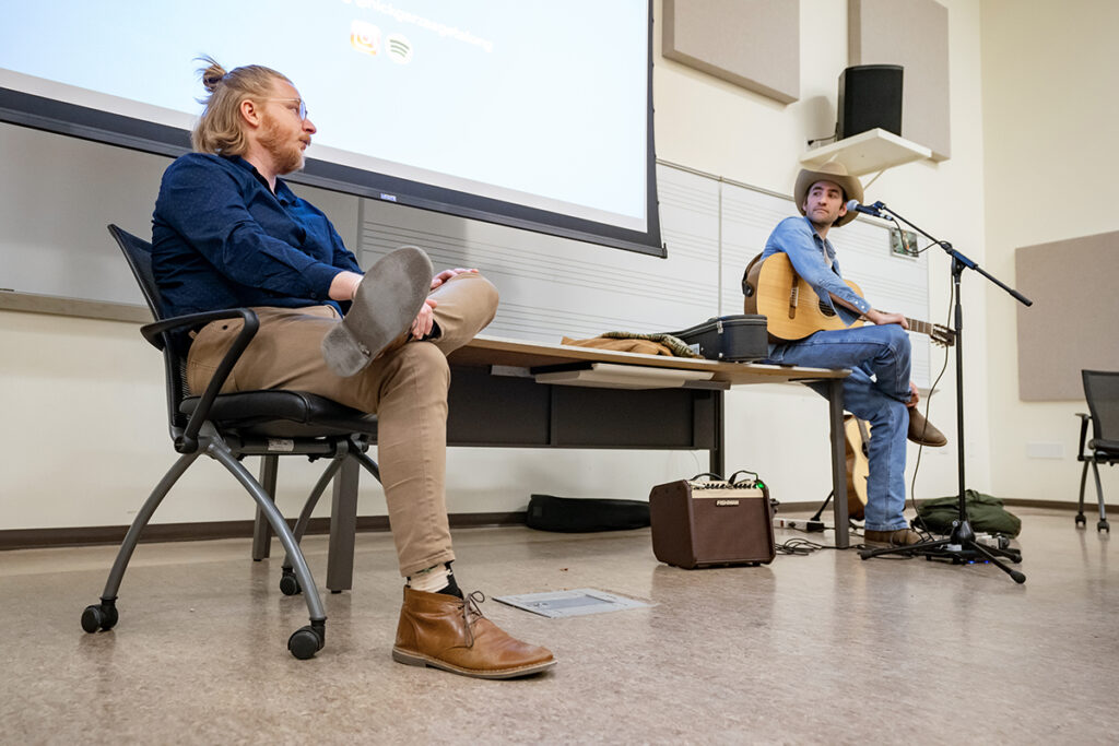 A country music performer and a college professor sit on chairs as they discuss music with college students in a classroom.
