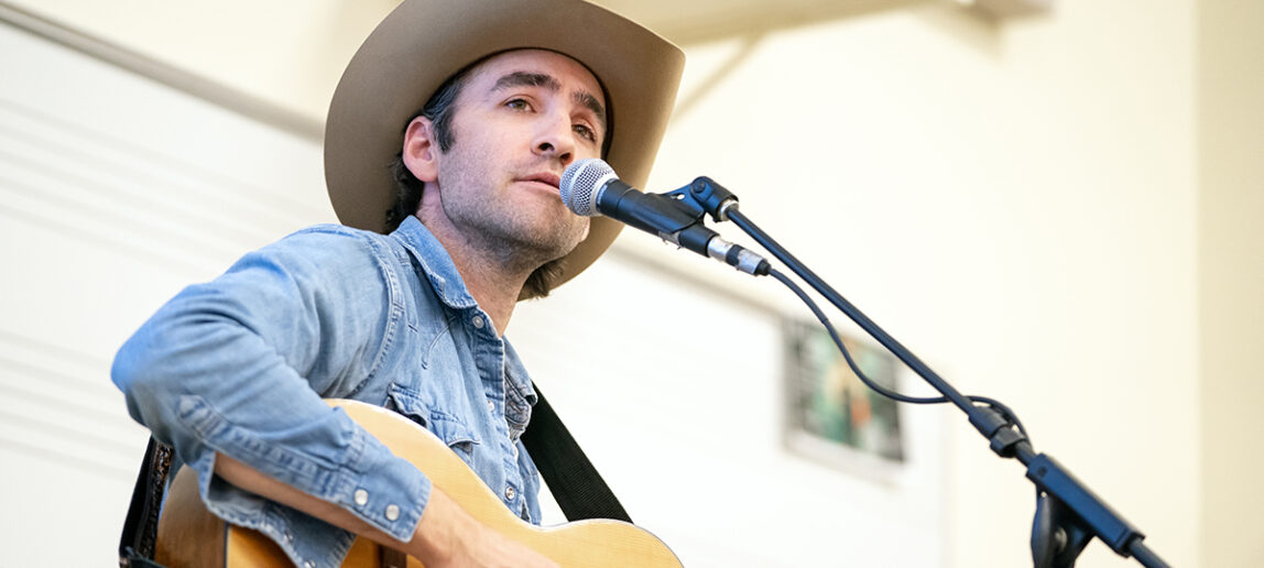 A country music performer sings and plays guitar as part of an event with college students in a classroom.