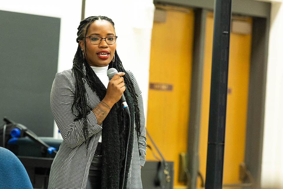 A performing artist speaks into a microphone as she addresses college students in a classroom setting.
