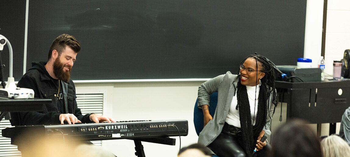 A performing artist smiles as she looks at a keyboard player seated to her right as they perform for college students in a classroom setting.