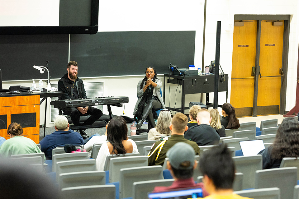A performing artist speaks into a microphone as she addresses college students in a classroom setting.