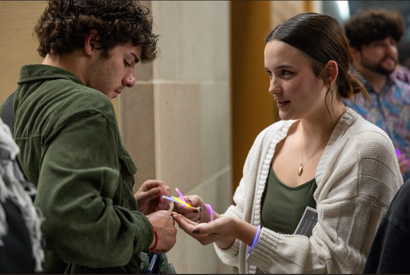 A college student helps another student put on a glow stick.