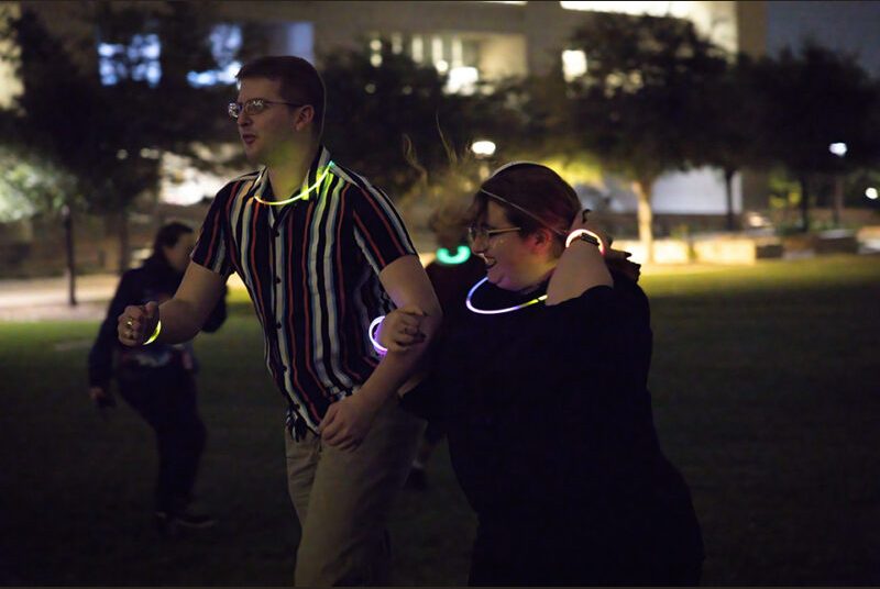 Two college students wearing glow sticks walk in an outdoor courtyard at night.