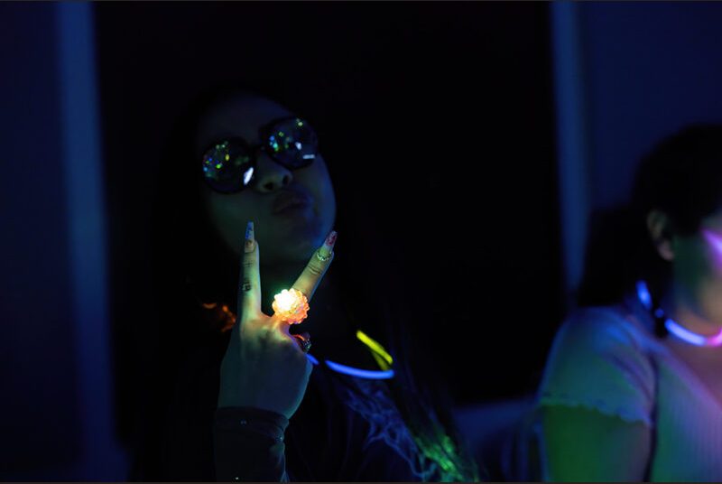 A college student wearing glasses and a glow stick gives the peace sign.