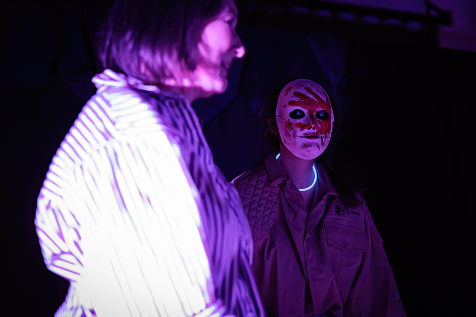 A person wearing a creepy white mask with a red handprint on it looks at a smiling woman.