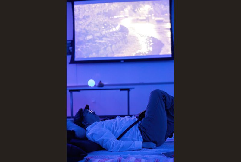 A person in a hat lays on a pillow and blanket, surrounded by blue light and a screen projecting an image.