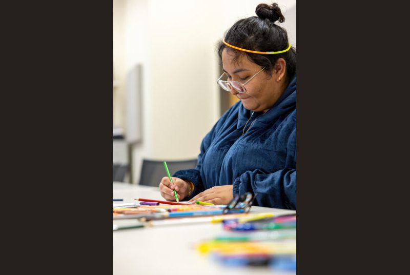 A college student sits at a table and draws using colored pencils.