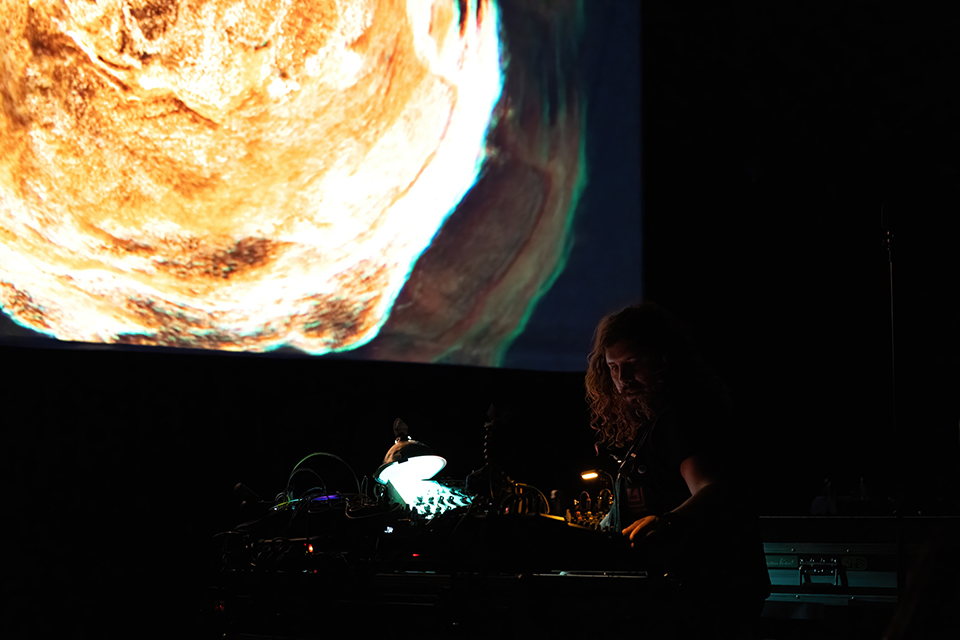 An artist performs with electronic equipment while a screen shows bright graphics behind him.