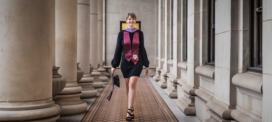 A college student walks through an ornate hall, wearing a black dress and a maroon Texas A&M sash and holding a graduation cap.
