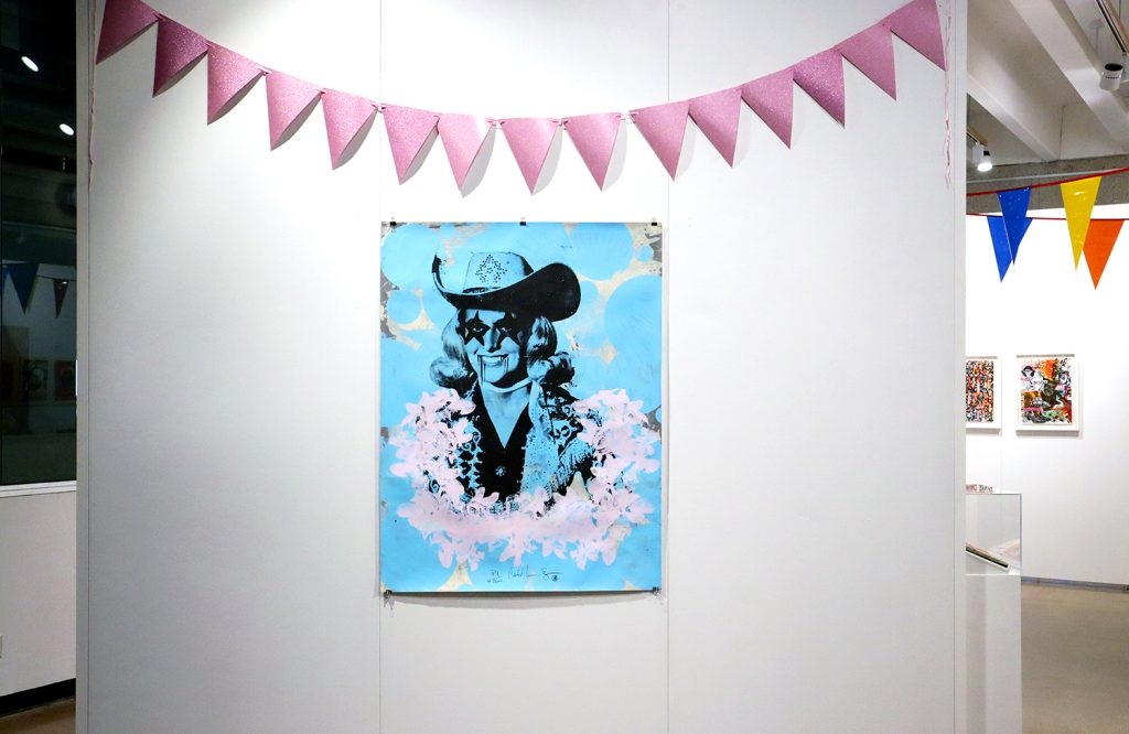 A blue art print hangs in a gallery, underneath a pink streamer ribbon. The image is of a woman wearing a cowboy hat, with stars over her eyes and dark lines coming from the edges of her mouth.