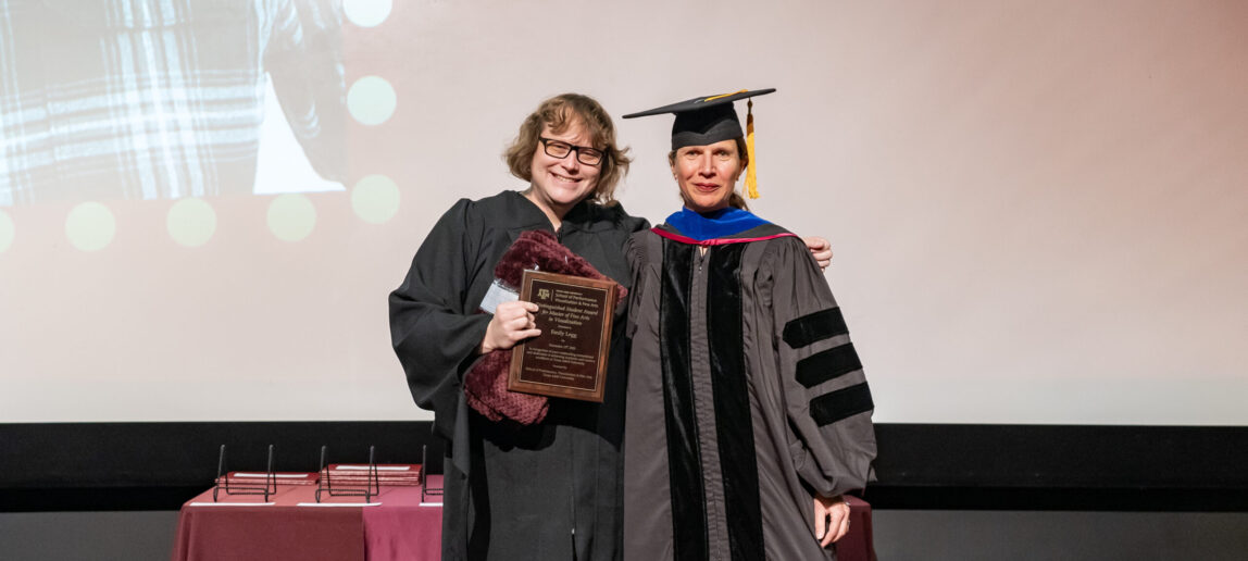 A college graduate student wearing a black graduation gown holds a plaque while standing by a professor wearing a black graduation cap and gown. Both are posing for a picture.