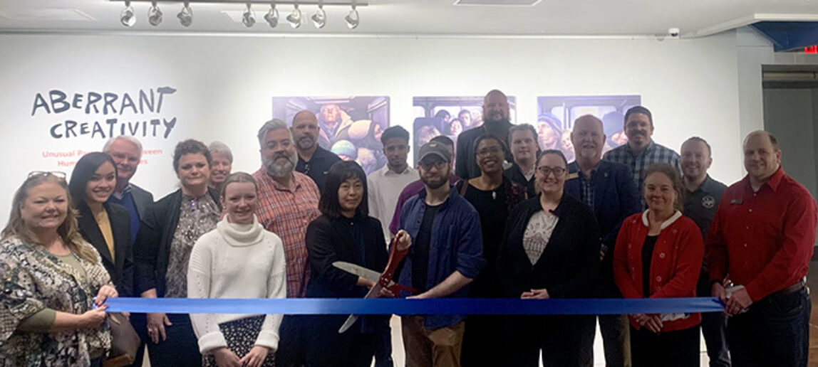 People gather for a ribbon cutting at an art exhibition, with two people holding oversized scissors by the ribbon.