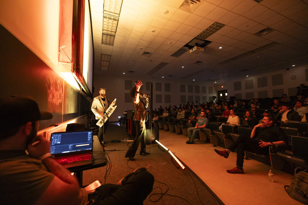 Two members of a band, one singing, one playing keyboards, perform for college students in a classroom. A lighting director is seen on the left hand side, working with a laptop computer.