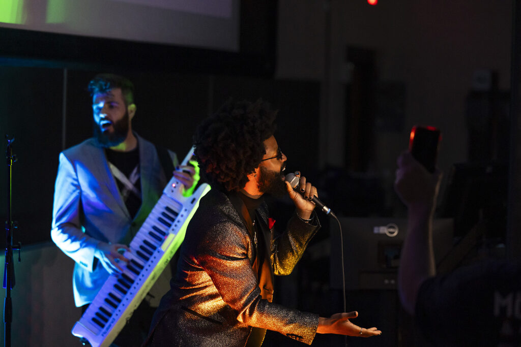 Two members of a band, one singing, one playing keyboards, perform for college students in a classroom.