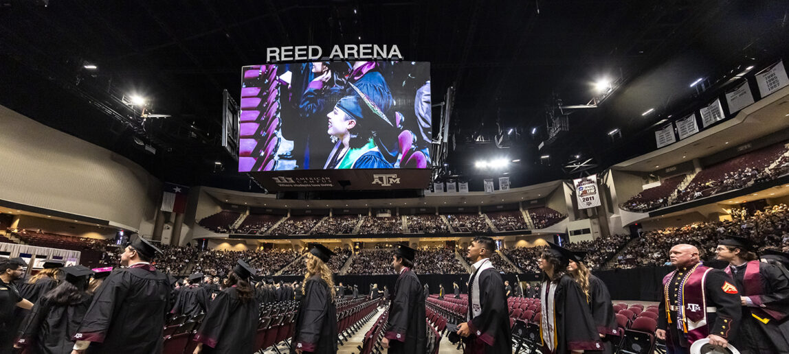 Graduating college students wearing caps, gowns and sashes file into a basketball arena for a graduation ceremony.