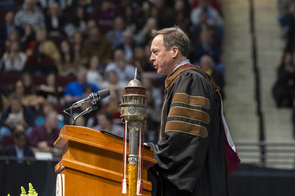 A university dean wearing a graduation robe speaks at a podium during a graduation ceremony.