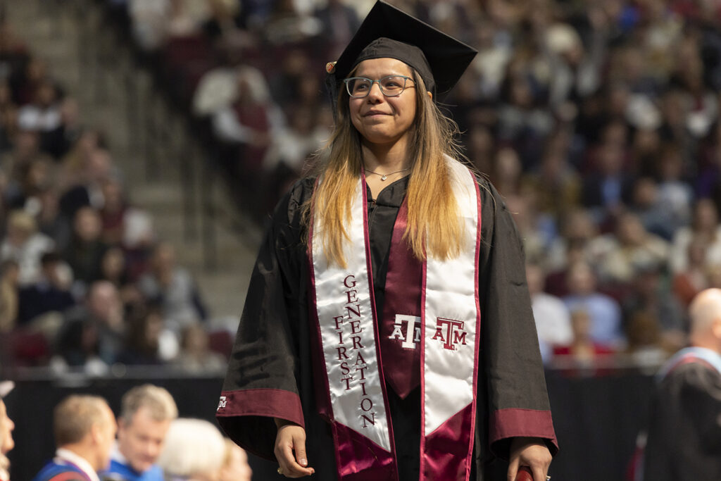 A college student in a cap and gown with a Texas A&M sash that says "First Generation" smiles as she walks across the stage at a graduation ceremony.