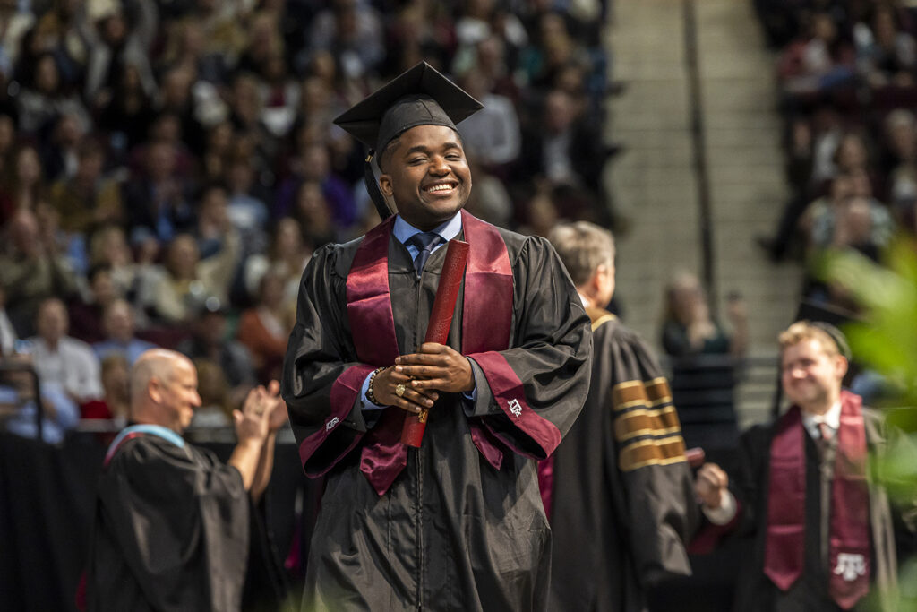 A college student in a cap and gown with a maroon Texas A&M sash holds his diploma tube and smiles as he walks across the stage at a graduation ceremony.