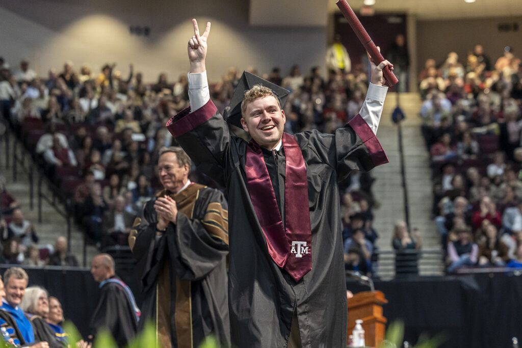 A college student walks the stage at a graduation ceremony, wearing a cap and gown and a maroon Texas A&M sash. He holds both hands up, one holding a diploma tube and the other making a peace sign.
