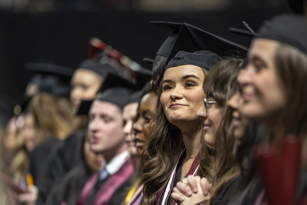 A graduating college student smiles as she sits among fellow students at a graduation ceremony.