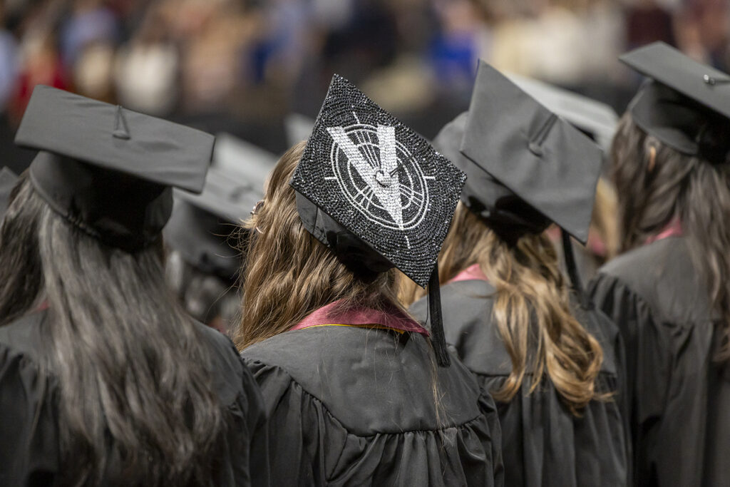 The back view of a college student's graduation mortar board, which is decorated in black and silver with a "V" graphic.