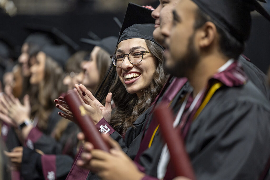 A college student sits among her classmates at a graduation ceremony, smiling and clapping.