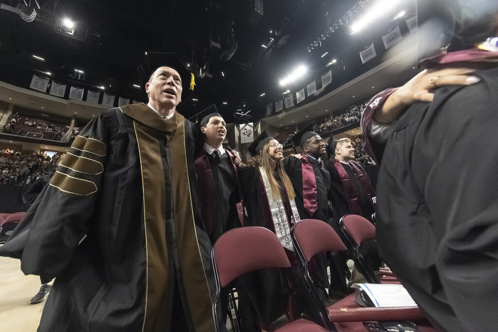 A university dean and four graduating students in caps and gowns lock arms and sway during the "Aggie War Hymn" song at a graduation ceremony.