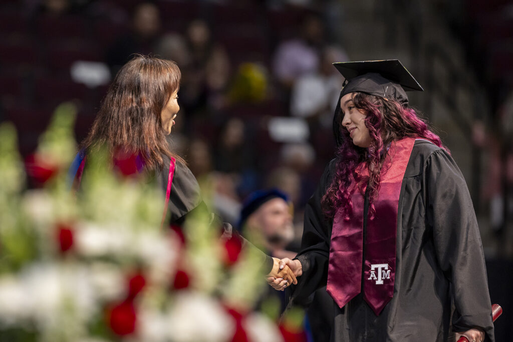 A student in a cap and gown and a maroon Texas A&M sash shakes the hand of the university official who handed her a master's degree diploma tube at a graduation ceremony.