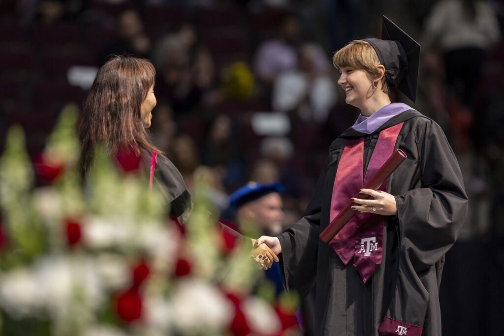 A student wearing a cap and gown with a purple sash and a maroon Texas A&M sash shakes the hand of the university official who just handed her a master's degree diploma tube at a graduation ceremony.