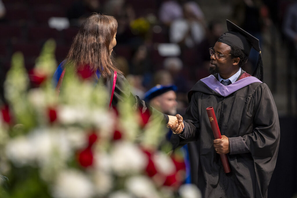 A student wearing a cap and gown with a purple sash shakes the hand of the university official who just handed him a master's degree diploma tube at a graduation ceremony.