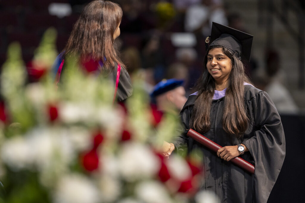 A student wearing a cap and gown with a purple sash shakes the hand of the university official who just handed her a master's degree diploma tube at a graduation ceremony.