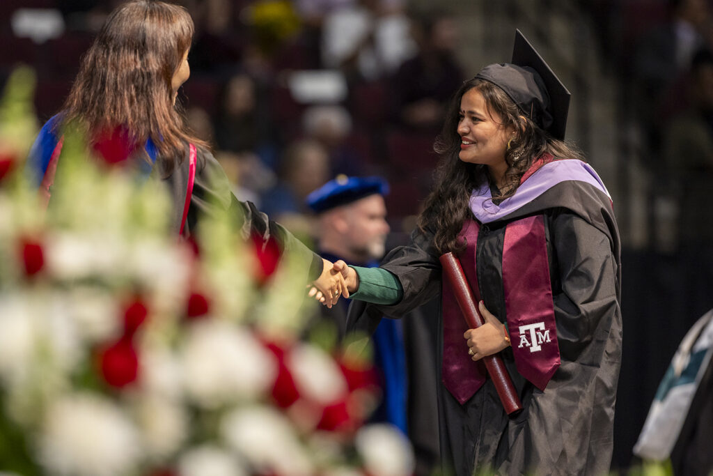 A student wearing a cap and gown with a purple sash and a maroon Texas A&M sash shakes the hand of the university official who just handed her a master's degree diploma tube at a graduation ceremony.