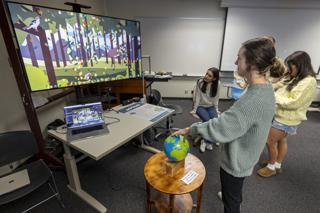 A college student touches a globe that is sitting on a table, which connects to a computer game about biomes.